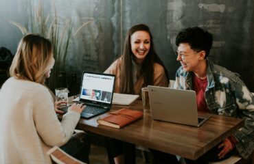 três estudantes usando notebook em cima de uma mesa, conversando e rindo